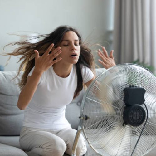 In living room without air-conditioner tired from summer heat young woman turned on floor ventilator waving her hands to cool herself, female sitting on couch suffers from unbearable too hot weather